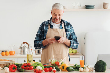 Online chat, dinner invitation, recipe app and technology at home. Smiling senior man in apron writes message while cooking salad in kitchen with table with many fresh vegetables
