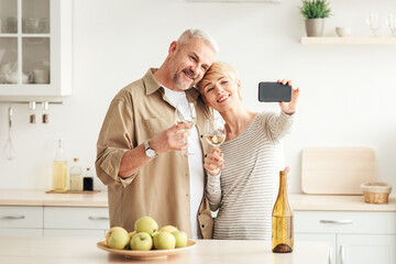 Selfie for holiday, app for phone, video call to family during covid-19. Cheerful middle-aged woman and man with wine on cozy kitchen interior, takes photo on smartphone with empty screen, free space