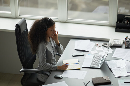 Young adult woman sitting at desk using headset while talking on phone, writing in notebook, working with laptop and documents, office environment with large windows