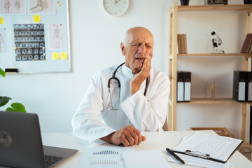 mature doctor with stethoscope works on a laptop looking at camera suffering from toothache, tooth deca. dental care concept. Senior therapist making video call, practitioner gp consulting