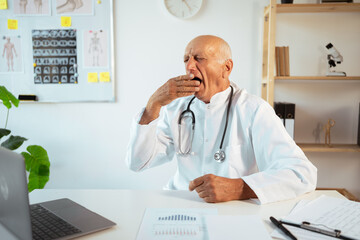 Tired mature doctor with stethoscope working overtime yawns, covers his mouth works on a laptop, looking at camera. Senior therapist making video call, practitioner gp consulting patient