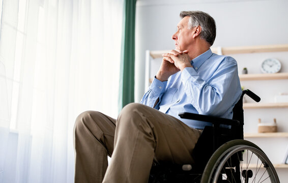 Low angle view of disabled senior male patient in wheelchair looking out window, feeling depressed at retirement home. Handicapped elderly man suffering from loneliness indoors