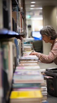 A librarian tidies up books on shelves near the circulation area multitasking between restocking and engaging with library guests requesting information.