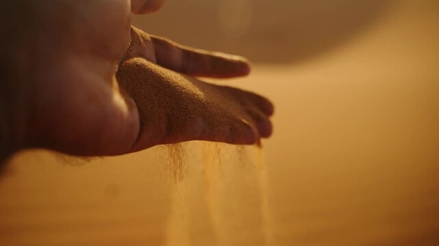Close-up of golden desert sand slipping gracefully through a hand at sunset in the Sahara Desert, Morocco, with warm light creating a soft glow.