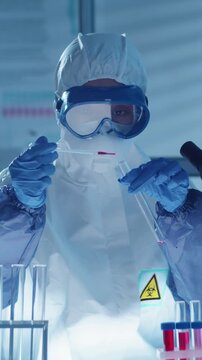 Vertical shot of scientist, wearing full personal prtective equipment, carefuly handling red liquid sampe in test tube with pipett in sterile laboratory