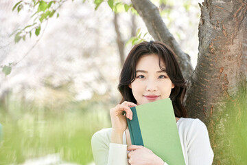 Young Asian female student holding a green book outdoors in spring park