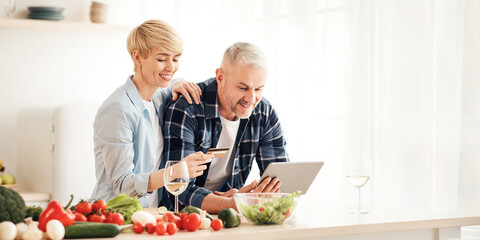Healthy eating in middle age, paying bills, ordering groceries and shopping online. Smiling senior couple use laptop and credit card in modern comfort kitchen interior with bright colored vegetables