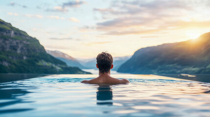 Man in serene infinity pool overlooks breathtaking mountain fjord, bathed in golden light of spectacular sunset, offering moment of peaceful natural immersion.