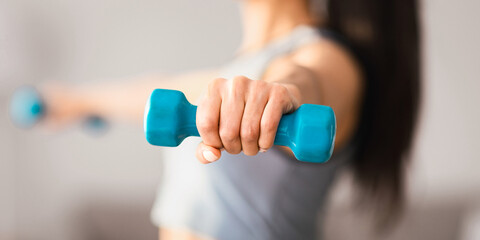 Selective focus on light blue dumbbell in unrecognizable sporty woman hand, closeup, blurred, copy space. Cropped of athletic lady training at home. Fitness, sport activity, healthy lifestyle