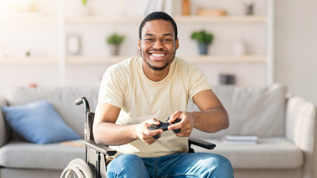Cheerful with disability man in wheelchair enjoying videogame on playstation, having fun at home. Gamer with joystick playing computer arcade. Hobbies concept
