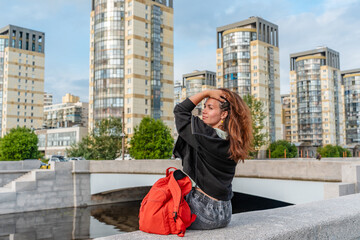 Fototapeta premium A young woman with backpack sits on the embankment in summer in evening city
