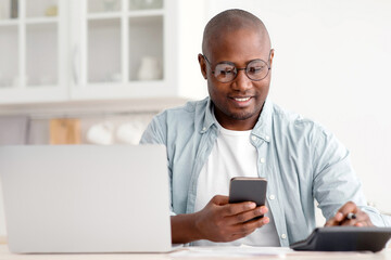 Savings and finances concept. Mature black man using calculator, phone and laptop computer, calculating taxes, sitting in kitchen at home, copy space