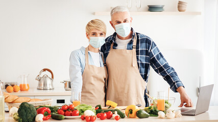 Covid-19 pandemic, healthy food blog and new recipes at home. Adult couple in aprons and protective masks in light kitchen interior with bright colored vegetables and laptop at morning, free space
