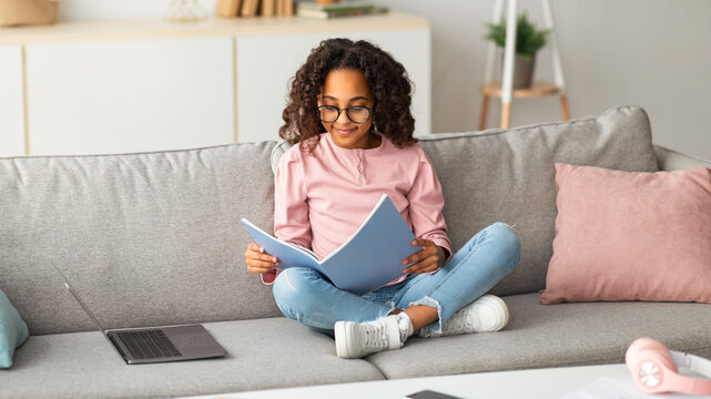 Learning and education concept. Smiling african american girl studying at home, sitting on sofa with laptop, reading and doing homework, holding notebook, copy space - Powered by Adobe