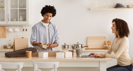 Remote work from home during COVID-19 pandemic. Millennial african american guy disassembles box of food from restaurant and gives to wife, female works behind laptop in kitchen interior, copy space