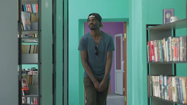 Young man in casual clothes dances joyfully in library hallway, wearing cap backwards and sunglasses hanging from shirt, moving with playful energy between bookshelves in bright modern interior