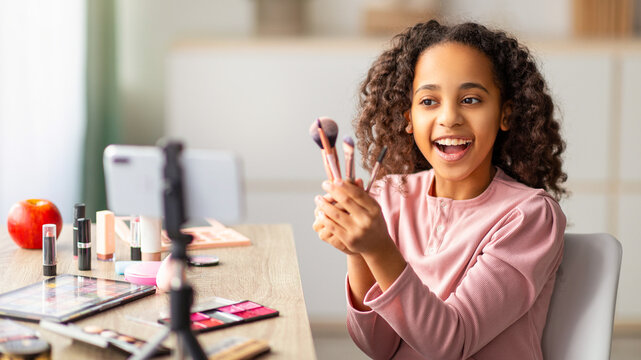 Young beauty blogger. Smiling girl showing her brushes, doing makeup and recording cosmetics product review. Teen looking at phone on tripod, filming video for her internet channel