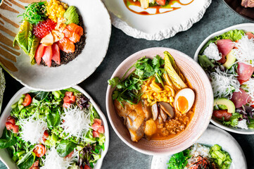 Assorted Asian dishes including ramen, poke bowls, and salads with fresh vegetables, seafood, and vibrant toppings arranged on a table