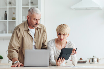 Problems with accounts, debts and mortgages, financial difficulties and tax. Serious sad mature european man and woman consider costs with laptop and calculator on kitchen interior, empty space