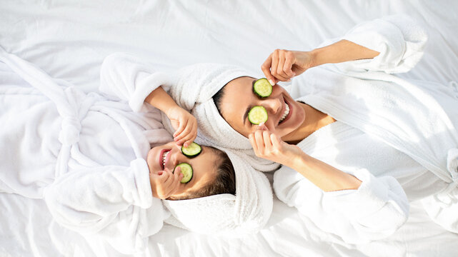 Top view of playful mother and daughter in bathrobes and white towels on heads laying on bed, holding cucumber circles, making face care procedures at home. Motherhood, parenthood concept - Powered by Adobe