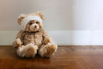 Obraz premium Teddy bear with white bandage on head sitting alone on wooden parquet floor.