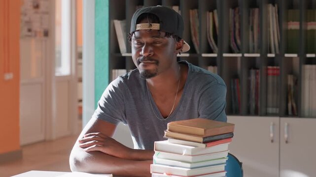 College student in cap sits at library table beside tall stack of books, arms resting and gazing ahead, ready for study session under warm light, shelves with magazines behind