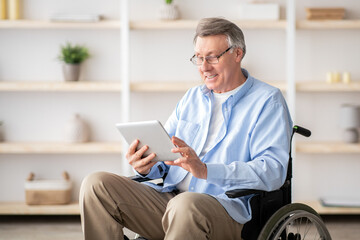 Joyful impaired senior man in wheelchair using tablet computer at home. Positive retired male with physical disability browsing web on touch pad, watching video online indoors