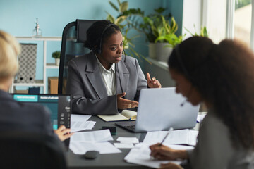 Black young adult woman wearing headset leading business meeting in modern office, gesturing while speaking to client, diverse coworkers listening and taking notes around table