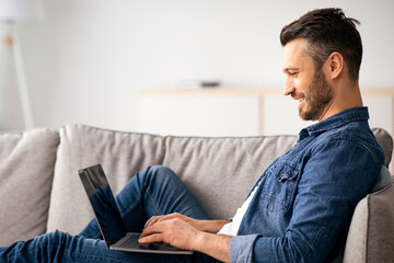 Side view of middle-aged man reclining on couch in living room, using newest laptop, copy space. Happy bearded man having fun at home, checking social media or chatting with friends on computer