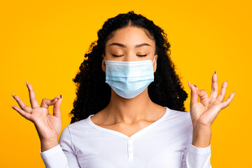 Dont Panic. Young black woman wearing medical mask meditating with closed eyes, feeling zen, millennial african american woman coping with pandemic stress, practicing yoga over yellow background