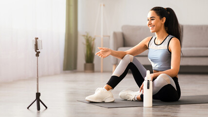 Pretty smiling young woman in sportswear sitting on yoga mat with bottle of water, having video...