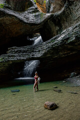 A young Caucasian woman stands in a shallow pool of water beneath a rocky overhang with a waterfall cascading in the background. The scene captures natural beauty in the Caucasus region.