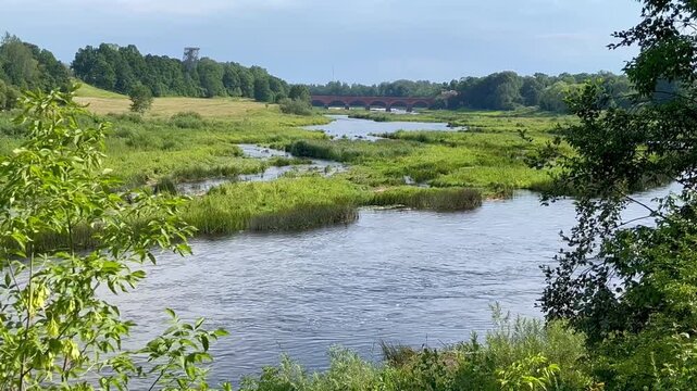 River Venta Flowing Past Green Marshlands Toward Iconic Red Arch Bridge in Latvia