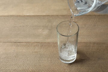 Pouring water from jug into glass at wooden table, closeup. Drinking clear water in the morning for health. Space for text 