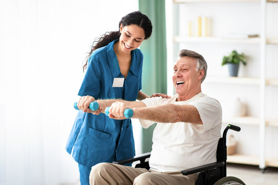 Young physiotherapist helping senior man in wheelchair do exercises with dumbbells, recovering from illness or injury at health centre. Rehab fitness for elderly patients concept - Powered by Adobe