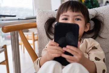 A young girl with pigtails smiles while using a smartphone, sitting comfortably on a chair indoors.