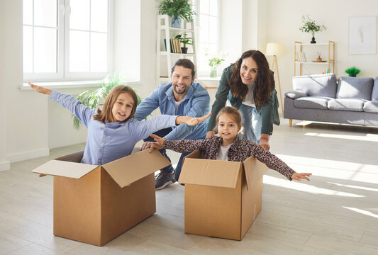 Joyful family having fun in new apartment as children pretending to fly in cardboard boxes while parents pushing them. Cheerful father, mother and kids celebrating purchase of new home and relocation.