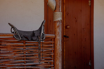 A rustic wooden house in the Caucasus region. The house features a woven fence and a saddle hanging on the porch. The window is partially open, revealing a glimpse of the interior.