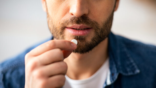 Treatment, medicine, medication, pills, vitamins, supplements concept. Cropped of bearded man taking white pill. Closeup of unrecognizable man holding medicine next to his mouth - Powered by Adobe