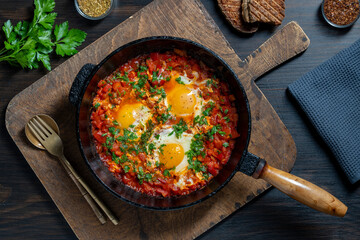 Shakshuka in frying pan for breakfast, closeup. Homemade shakshuka from poached eggs, onion, bell pepper, olive oil, red tomato and parsley in a cast iron skillet, top view. Mediterranean cuisine