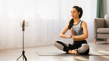 Fototapeta premium Yoga class, meditation course online concept. Sporty woman yoga teacher sitting on fitness mat with closed eyes in lotus pose in living room and meditating, broadcasting from home, copy space