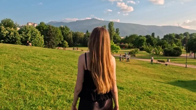 Sofia, Bulgaria - 16.06.2025. Relaxed woman walking in the city park on the green grass lawn towards the mountains. Weekend pleasure in nature. Wide angle landscape of park and sky with clouds.