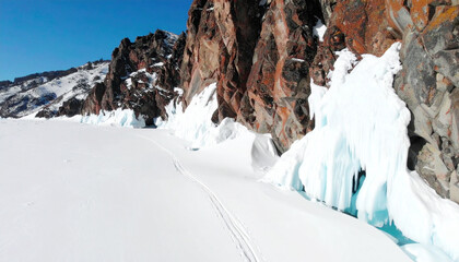 Frozen lake shore with icy cliffs and snow-covered terrain, showing tracks in the snow.