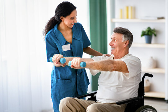 Female physiotherapist helping elderly man in wheelchair do exercises with dumbbells at health centre. Senior male petient undergoing rehabilitation after illness or injury at fitness clinic
