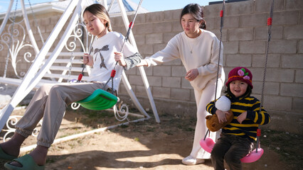An Asian woman rocks her daughters on a swing. A caring mother helps her two children swing on a swing on a sunny day
