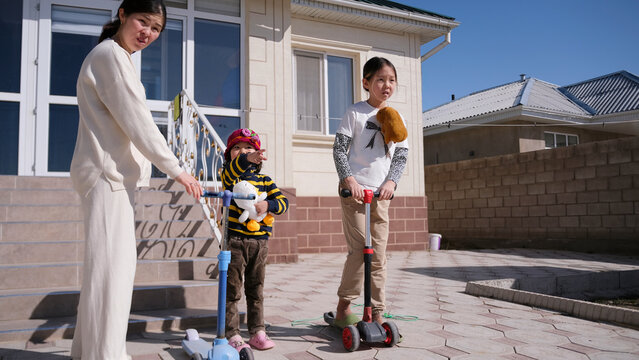 A typical Kyrgyz family. A woman and her two daughters went out into the yard to ride scooters. Funny kids spend time with their mom