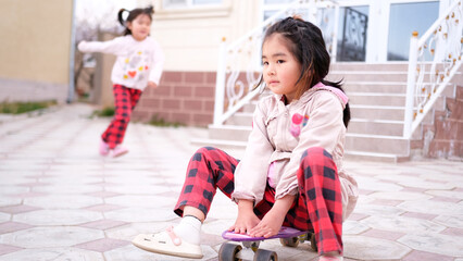 Two sisters are having fun in the courtyard of the house. An Asian girl in plaid trousers sits on a skateboard