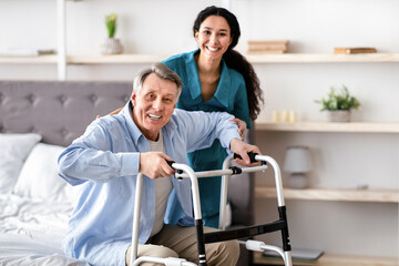Obraz premium Positive female nurse helping elderly male with walking frame stand up from bed at home. Senior man receiving assistance from medical worker indoors. Professional care for disabled patients