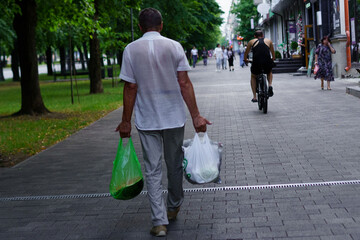 Older man walks along a park path carrying grocery bags while a cyclist passes by during the evening hours