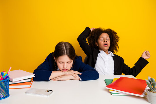 Two multicultural schoolgirls in a classroom setting, one yawning and the other resting her head, displaying school life moments
